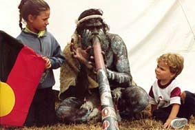 A picture of an indigenous man playing a didgeridoo with 2 children