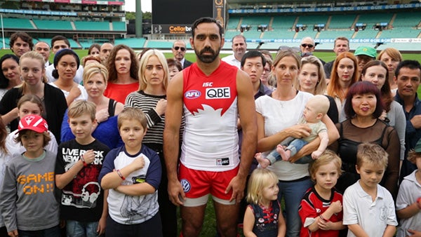 Adam Goodes in field. Group of people stand behind him in unison